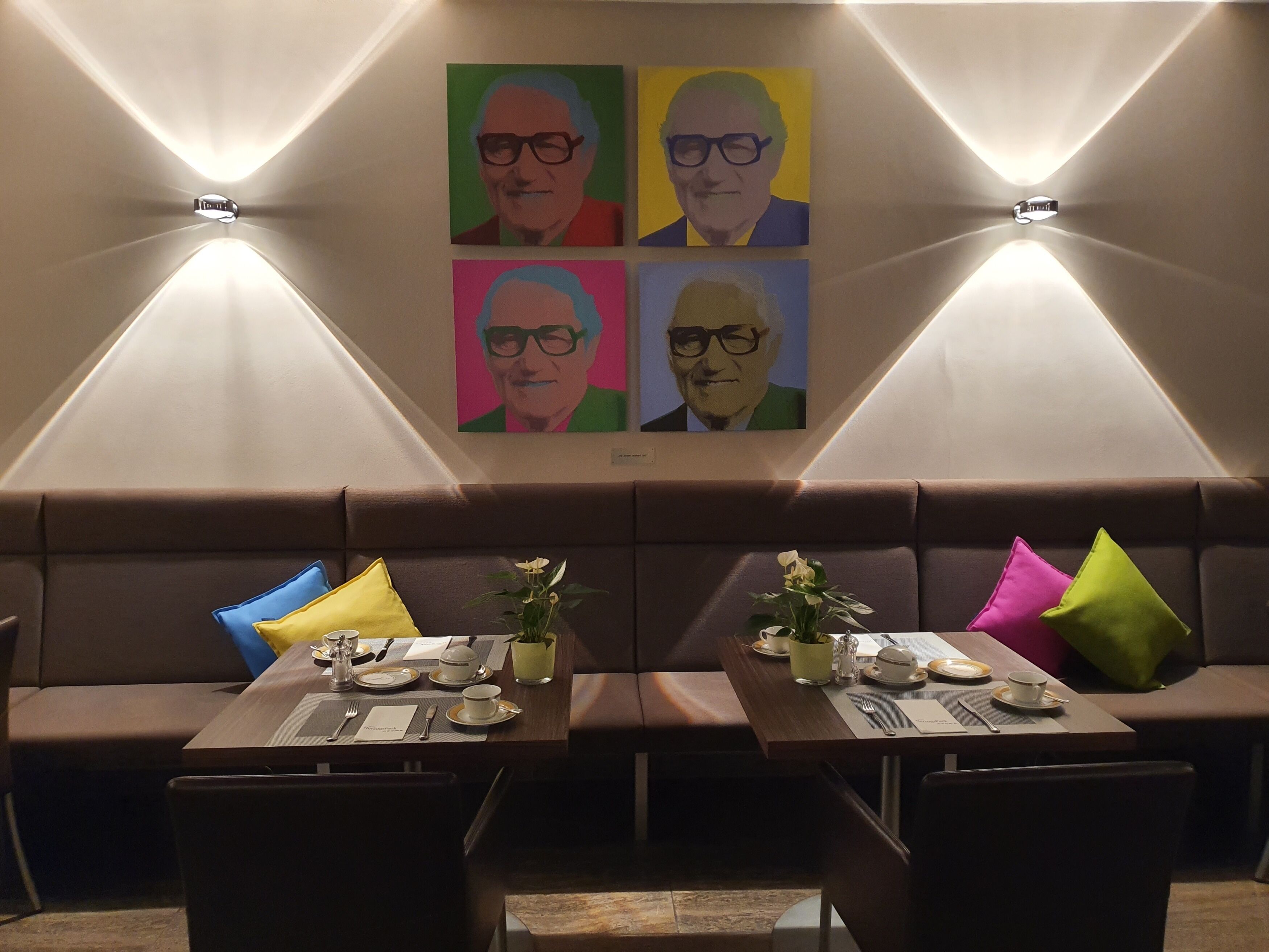 A dining area with two tables set for two, each with a vase of flowers, a menu, and cutlery. The walls feature colorful artwork.