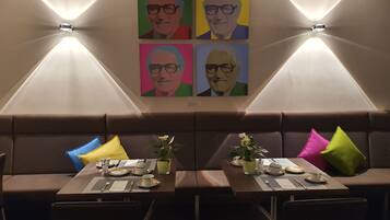 A dining area with two tables set for two, each with a vase of flowers, a menu, and cutlery. The walls feature colorful artwork.
