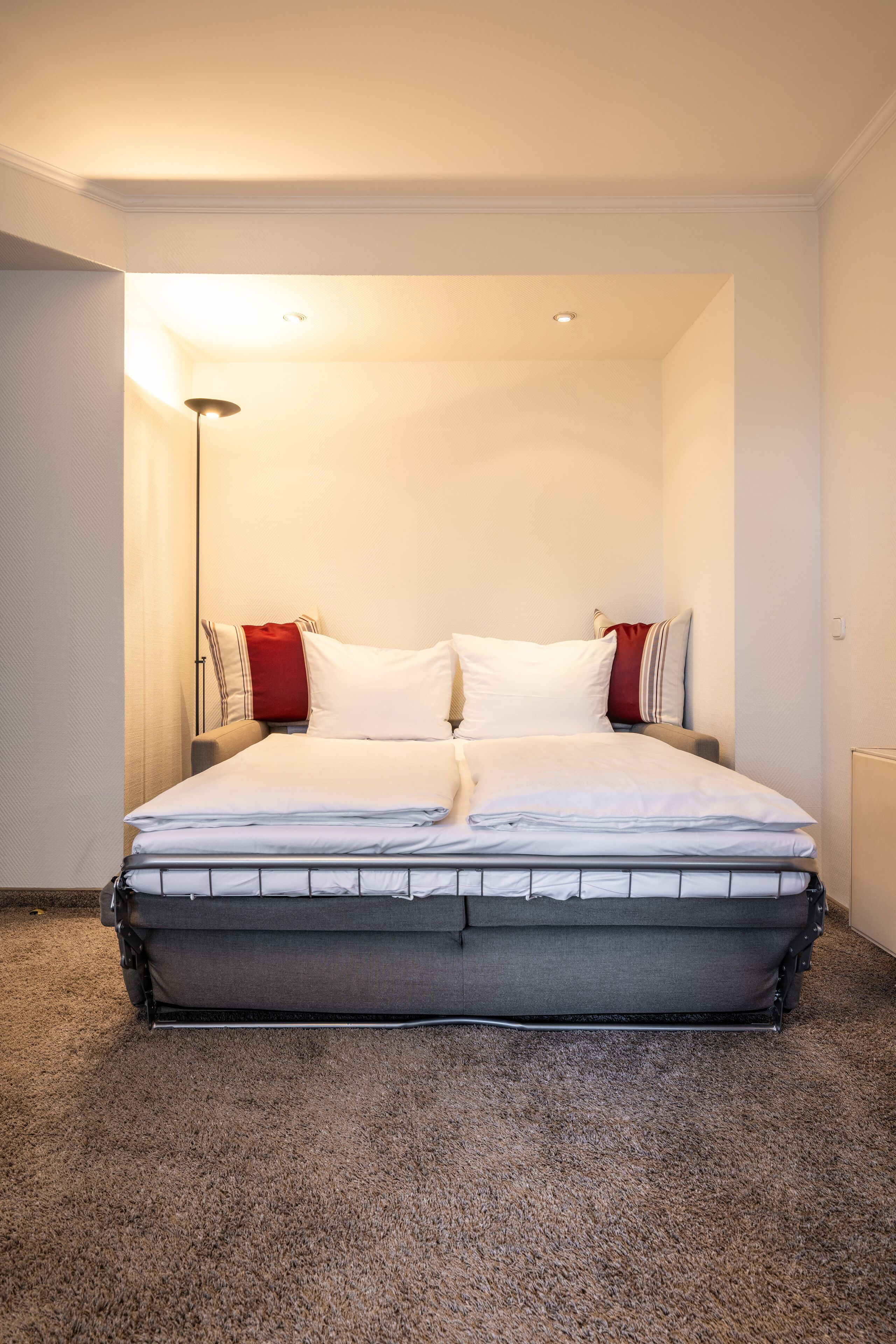 A neatly made bed with white and red pillows, a grey mattress, and a grey headboard.