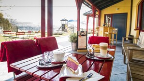 An outdoor café with wooden tables and red chairs, a view of a playground, and a covered seating area with a yellow wall and red accents. - JUFA Hotel Maria Lankowitz am See (Maria Lankowitz)