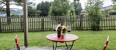 Un patio con una mesa metálica roja y dos sillas rojas, una botella de vino y un jarrón con flores.