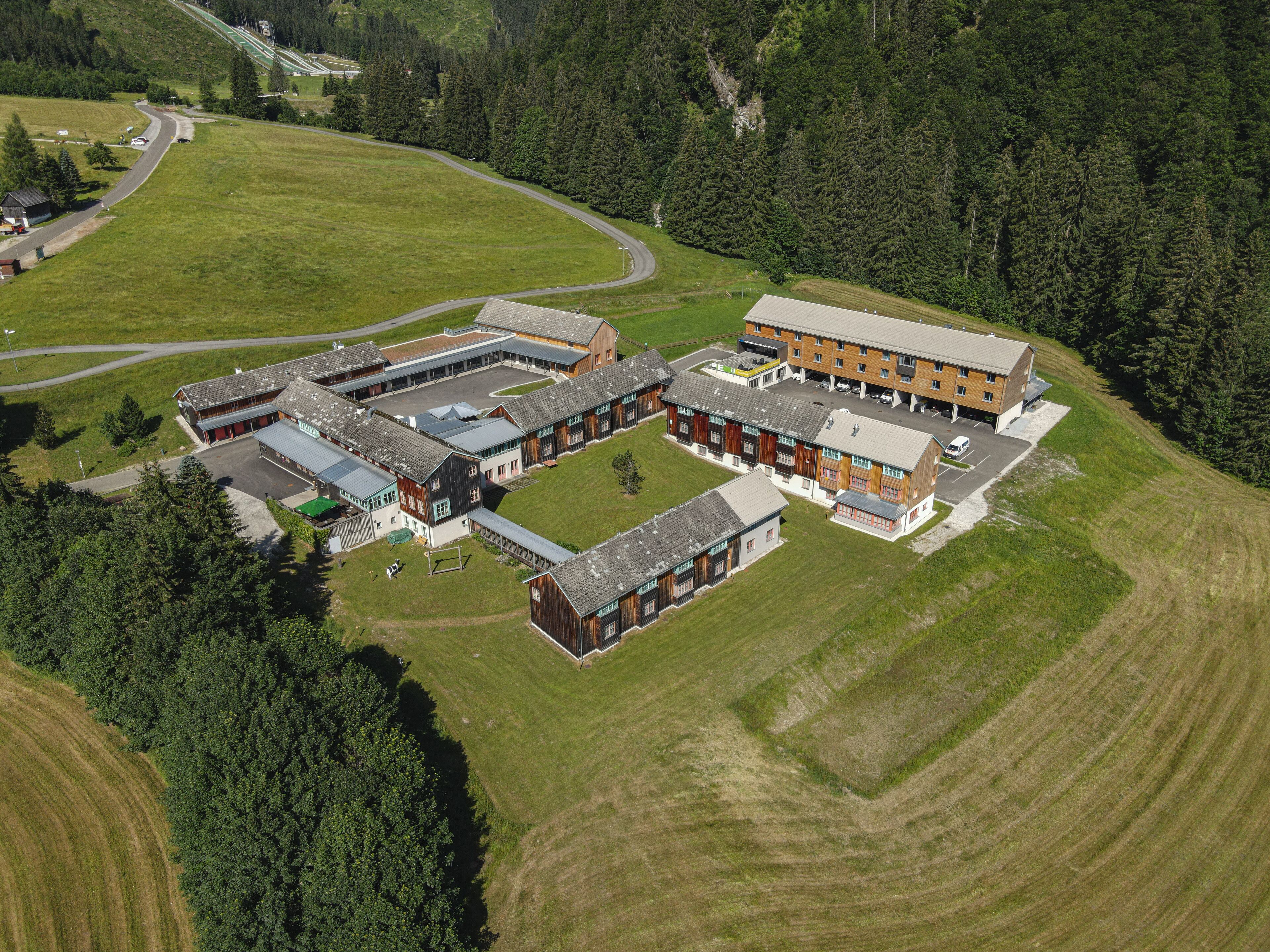 An aerial view of a modern building complex with multiple structures, surrounded by green fields and dense forest.