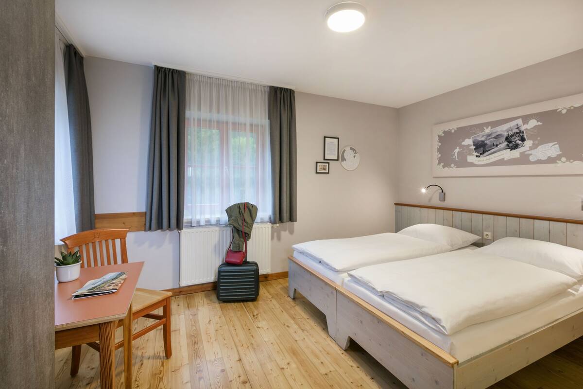 Een hotelkamer met een houten vloer, twee eenpersoonsbedden, een klein tafeltje met een stoel, een radiator en een raam met gordijnen.