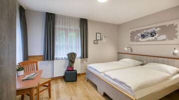 Habitación de hotel con piso de madera, dos camas individuales, una mesita con una silla, un radiador y una ventana con cortinas.