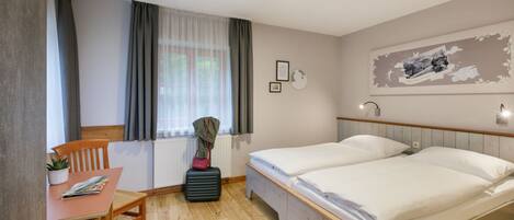A hotel room with a wooden floor, two single beds, a small table with a chair, a radiator, and a window with curtains.
