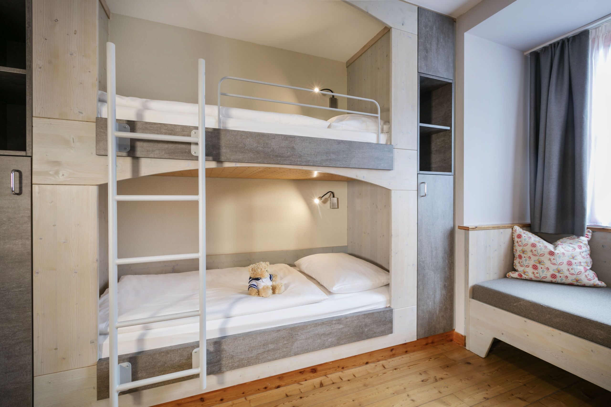 A bunk bed with a teddy bear on the lower bunk, a bedside table with a lamp, and a window with curtains.