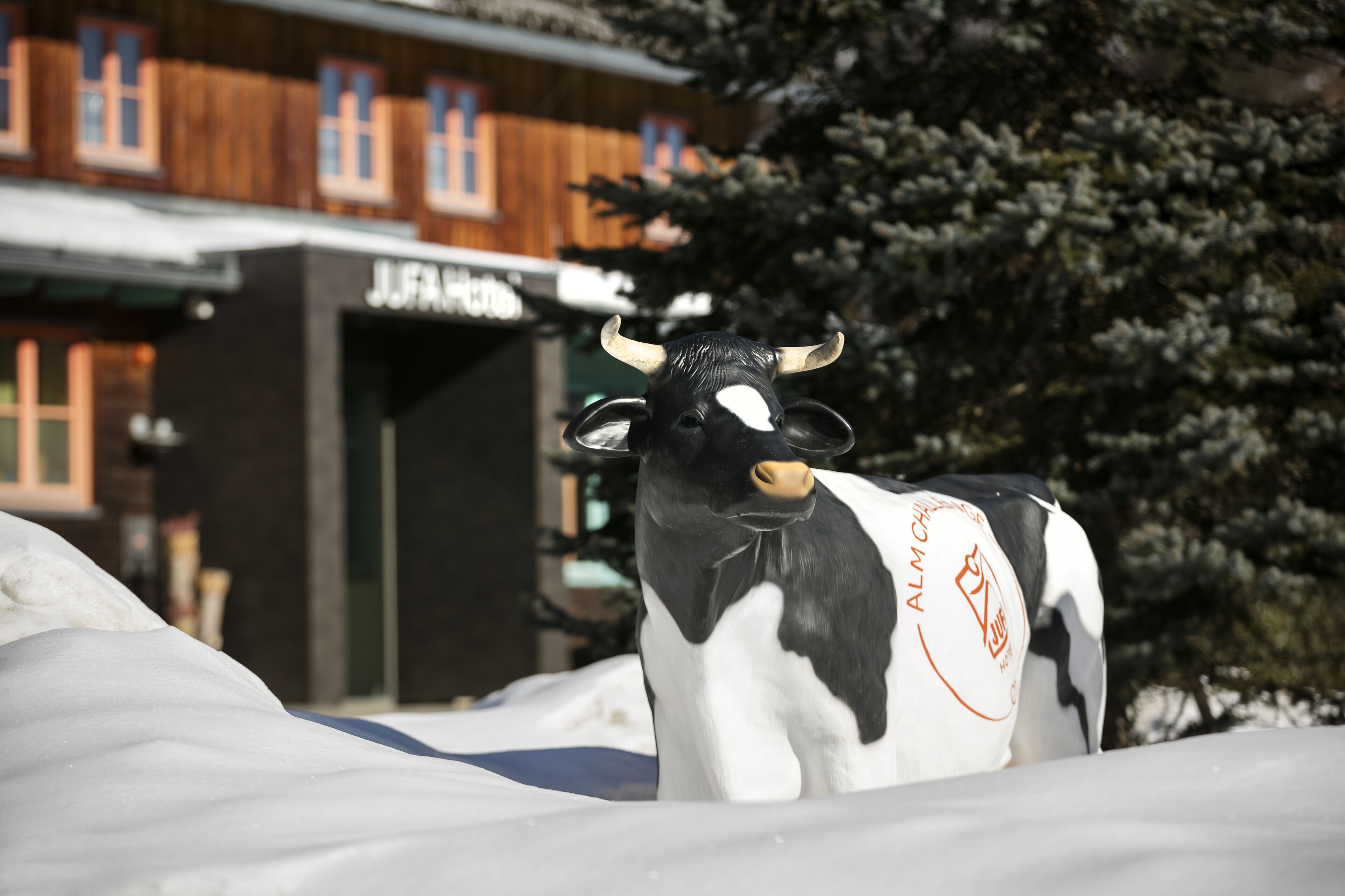 Paesaggio innevato con una statua di mucca in primo piano e un edificio con un cartello che riporta la scritta "JUFAR".