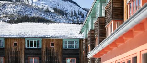 A row of wooden buildings with multiple windows and a snow-covered hill in the background.