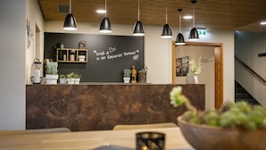 A reception area with a wooden ceiling, black pendant lights, a chalkboard with text, potted plants, and a wooden counter.