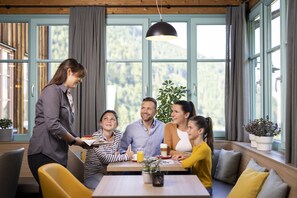 A family dining in a room with large windows, a wooden ceiling, and a table with a plant and a glass of juice. - JUFA Hotel Eisenerz (Eisenerz)
