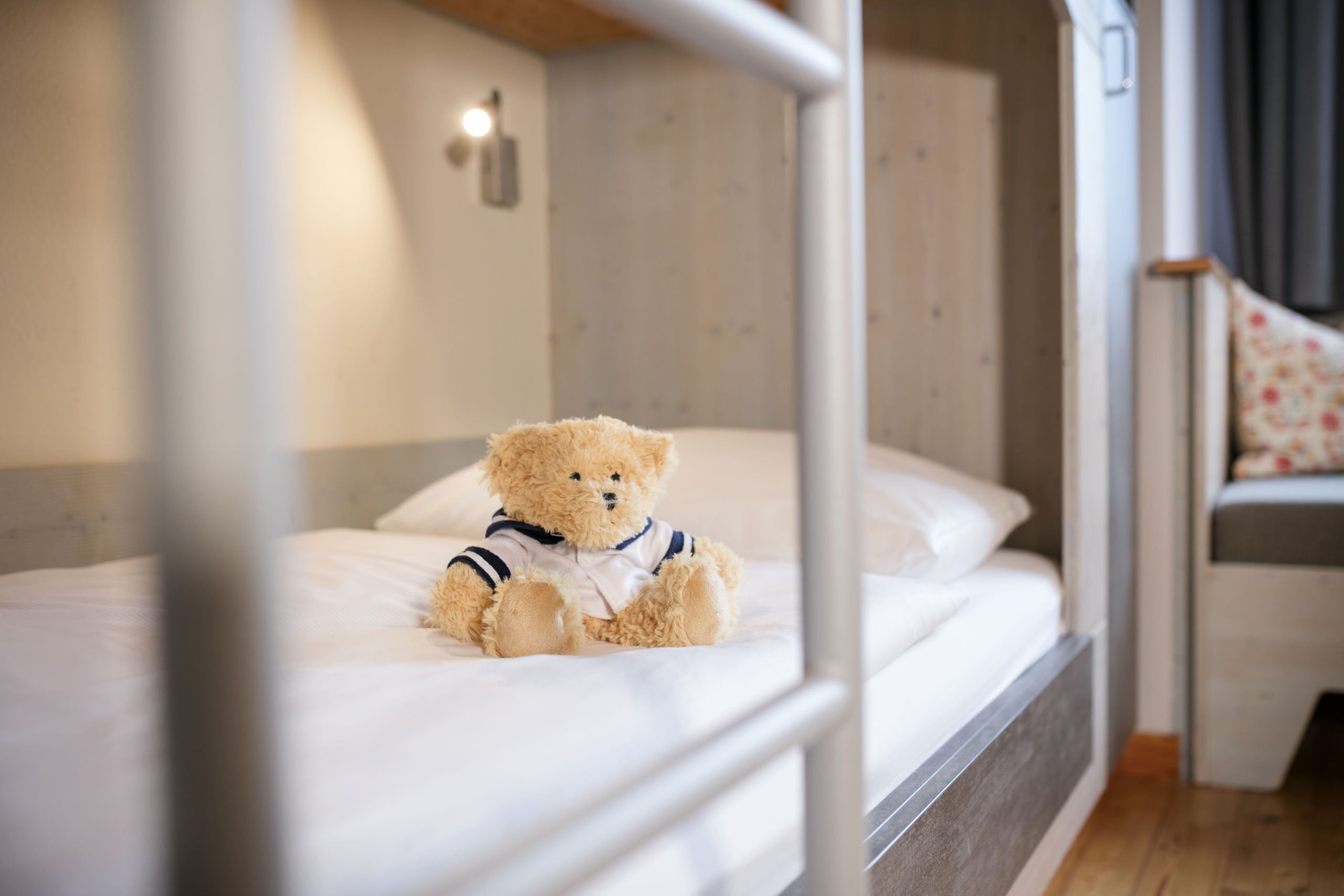 A single bed with a teddy bear on it, a bedside table, and a small chair with a floral cushion.
