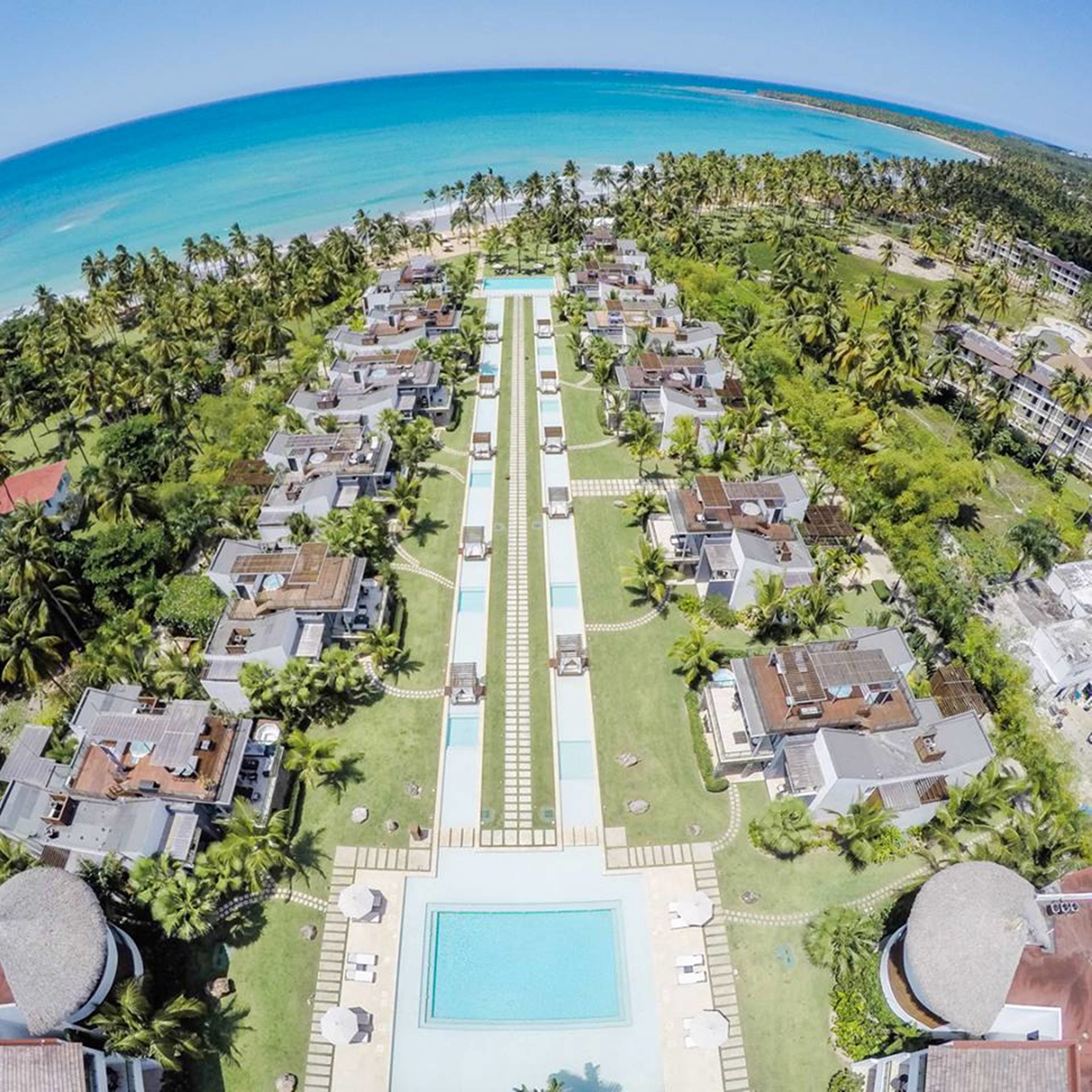 Luchtbeeld van een resort met een centraal zwembad, omringd door villa's en palmbomen, met uitzicht op een strand.