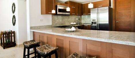 A modern kitchen with wooden cabinets, stainless steel appliances, and a granite countertop. There are three bar stools with woven seats.