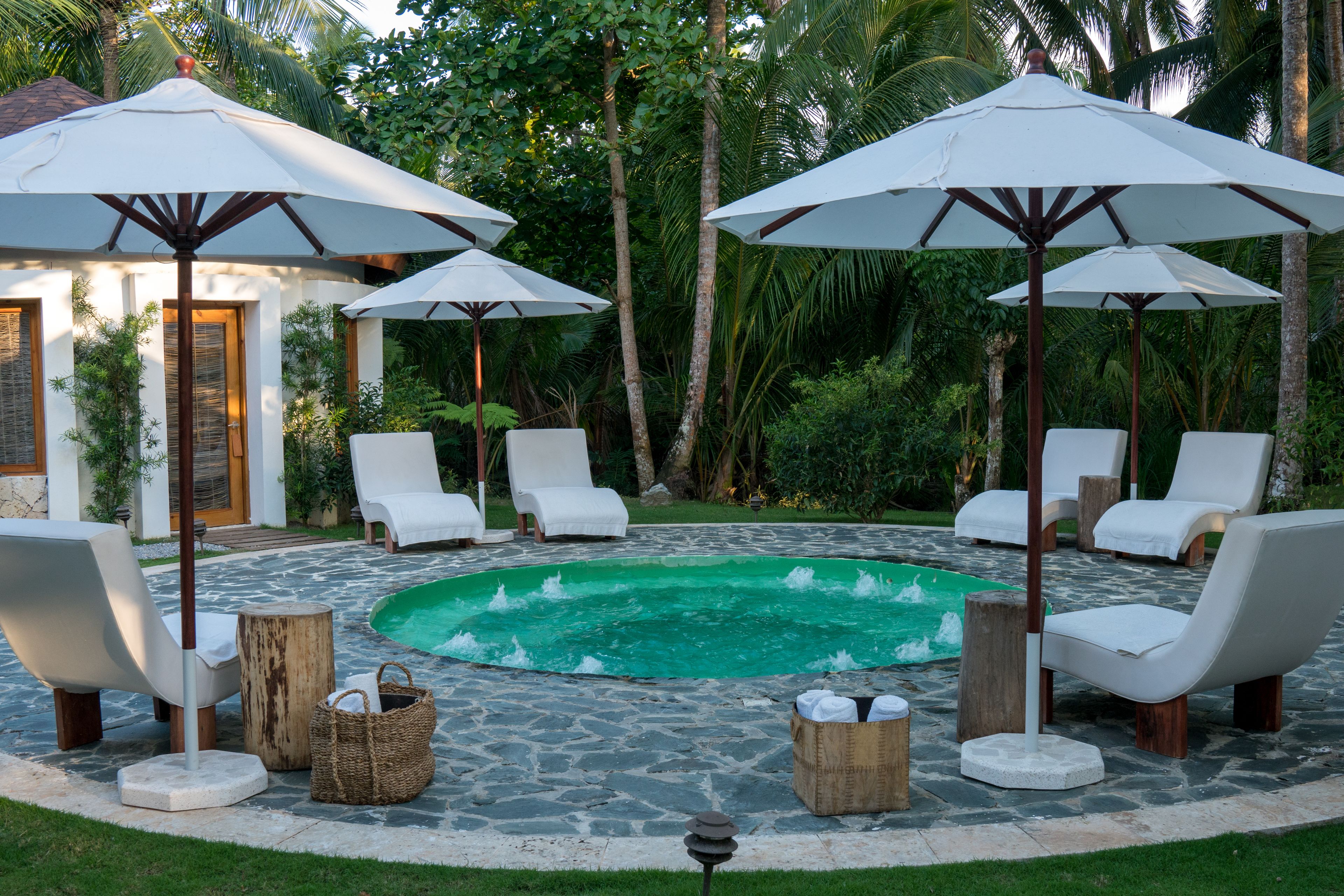 Un espace piscine agrémenté de chaises longues blanches et de parasols, entouré d’un sol en pierre et de plantes tropicales.