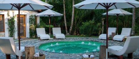 Un espace piscine agrémenté de chaises longues blanches et de parasols, entouré d’un sol en pierre et de plantes tropicales.