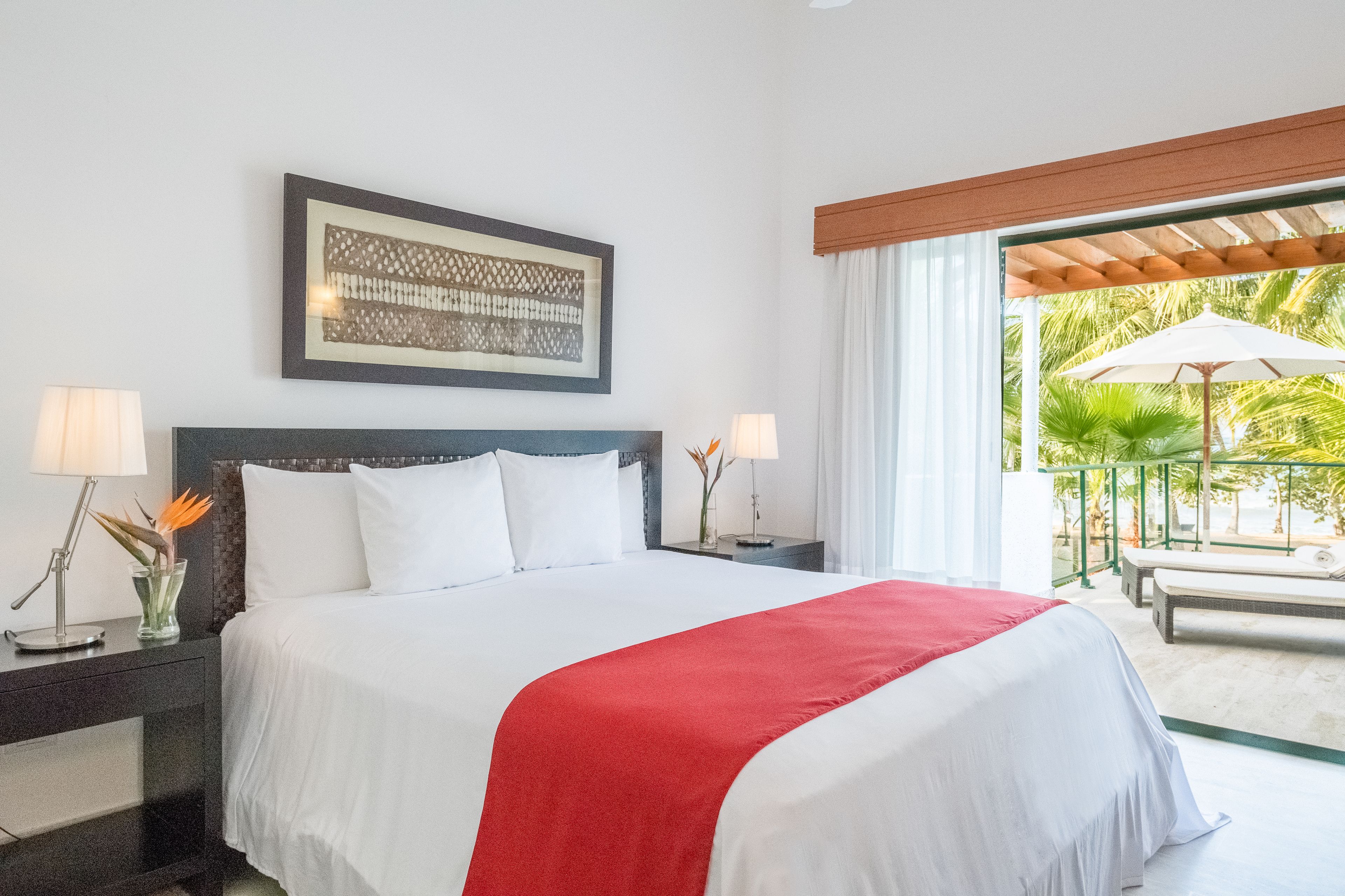 A hotel room with a large bed, two bedside tables with lamps, a framed artwork above the bed, and a view of a palm tree and outdoor seating area through a large window.