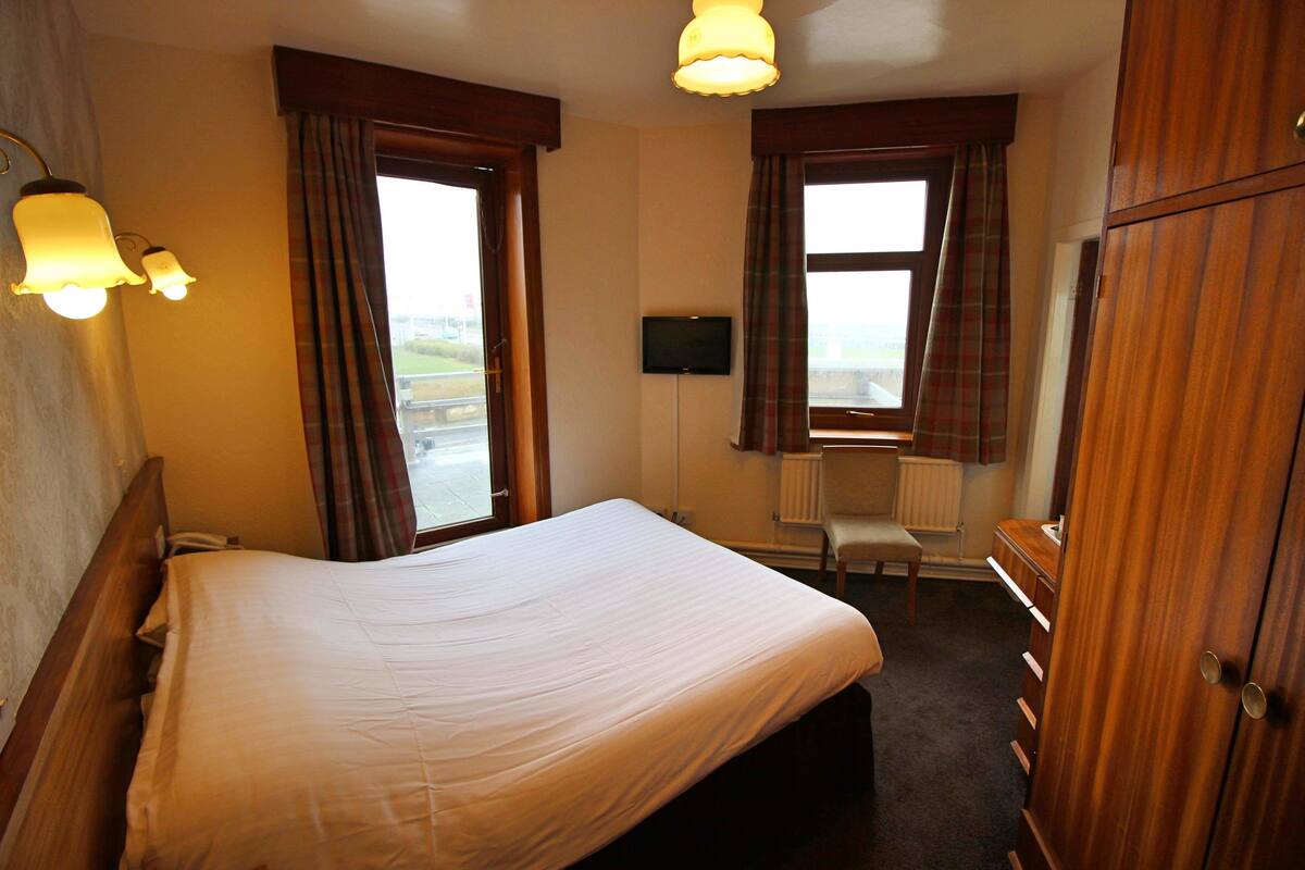 A hotel room with a large bed, two windows with curtains, a television on the wall, and a wooden wardrobe.