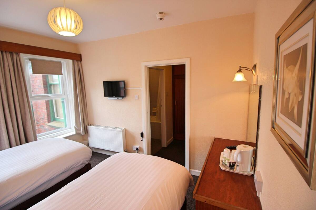 A hotel room with two beds, a television, a radiator, a bedside table with toiletries, and a framed picture on the wall.
