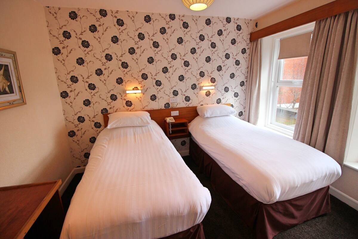 A hotel room with two single beds, floral wallpaper, bedside lamps, a wooden headboard, and a window with curtains.