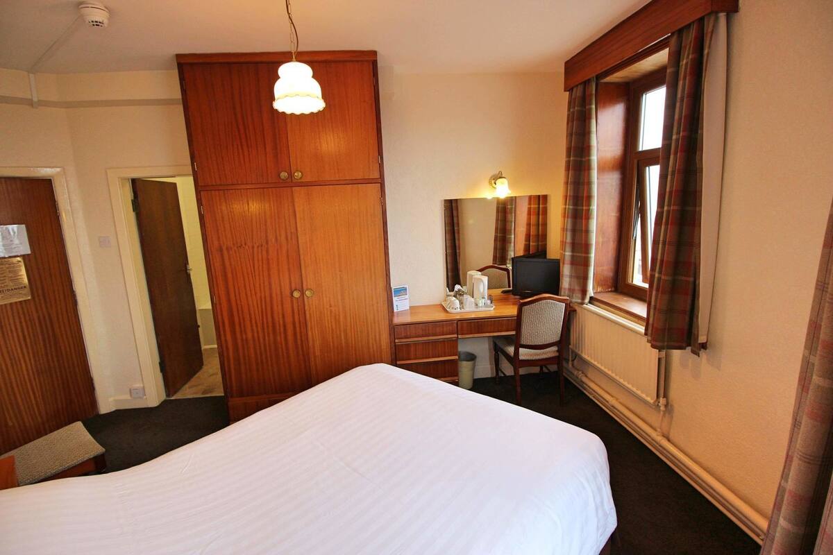 A hotel room with a bed, a desk with a chair, a wardrobe, a mirror, and a window with curtains.