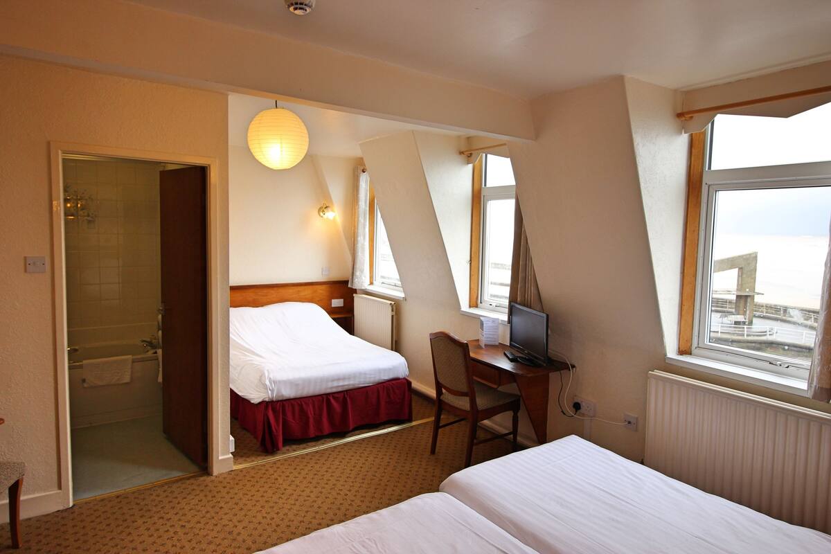 A hotel room with a bed, a desk with a computer, a chair, a window with a view, and a bathroom with a shower and toilet.