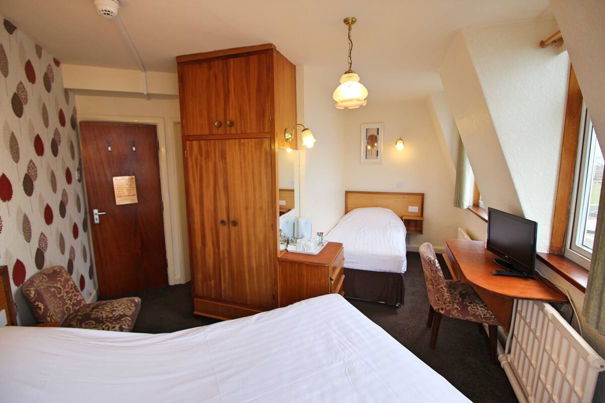 A hotel room with two beds, a wooden wardrobe, a desk with a computer, and a window with a view.