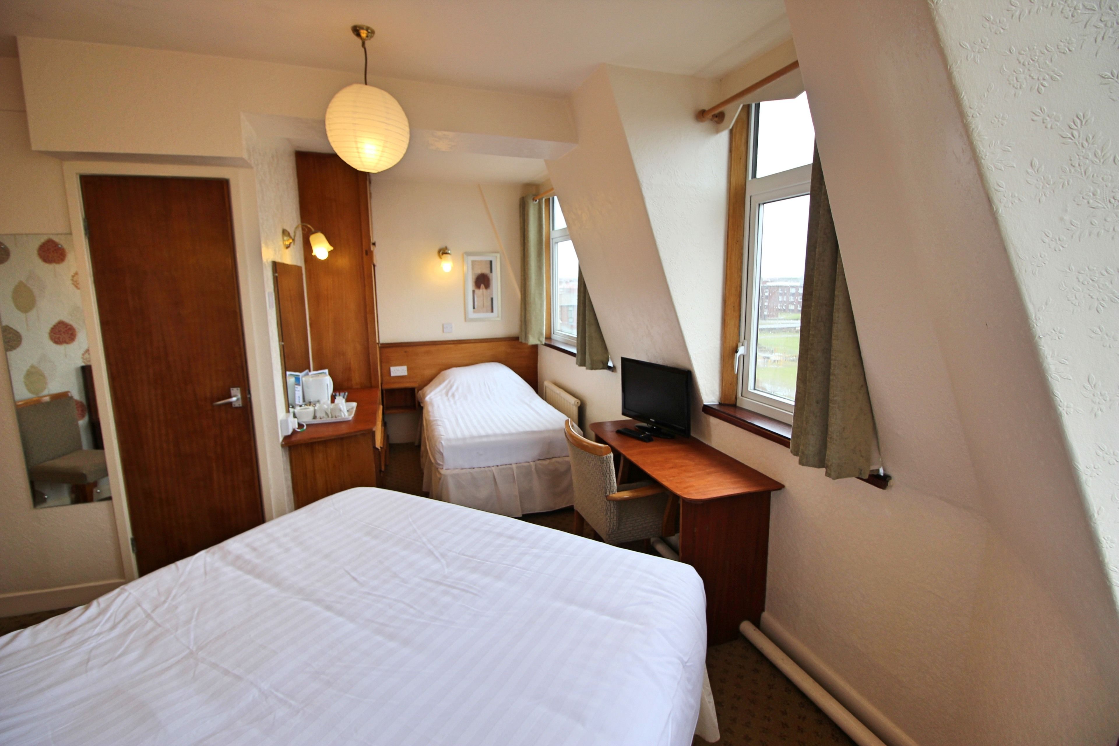 A hotel room with a large bed, a desk with a computer, a chair, a small table with a lamp, and a window with curtains.