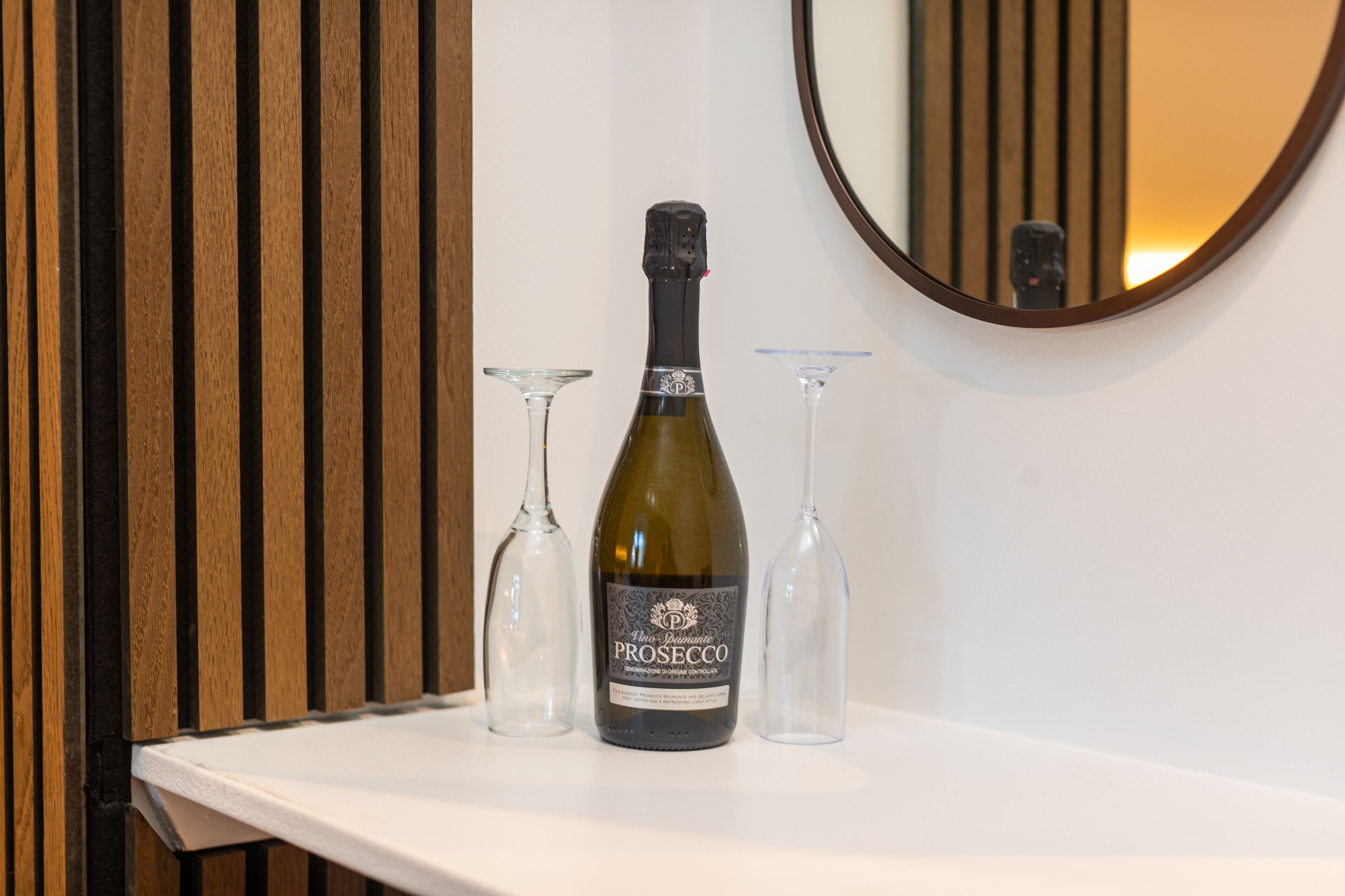 Una botella de vino Prosecco sobre una superficie blanca, con dos copas de vino vacías a su lado, frente a un panel de madera y un espejo.