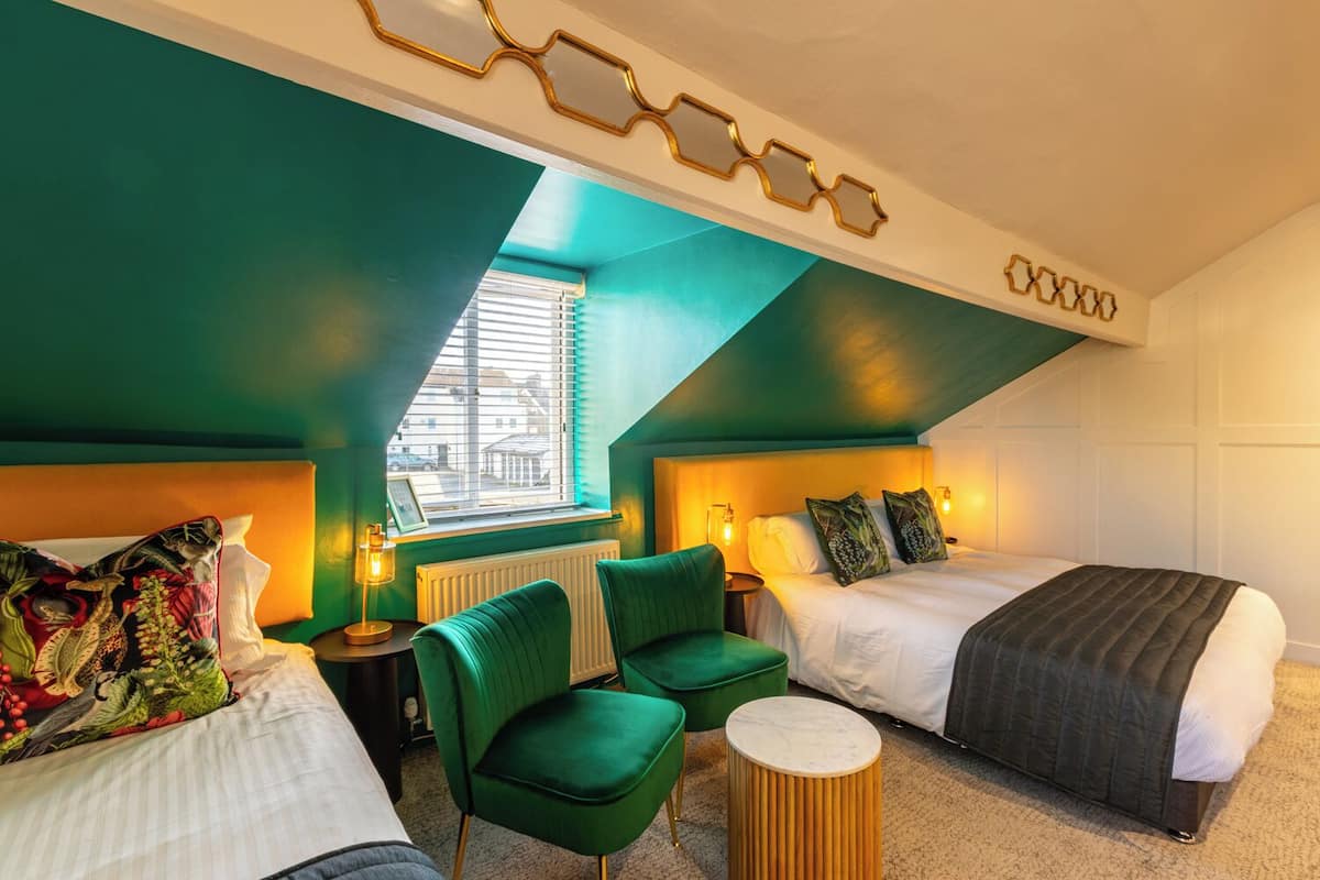 A modern hotel room with a sloped ceiling, green walls, two beds, a green armchair, a round side table, and a window with blinds.