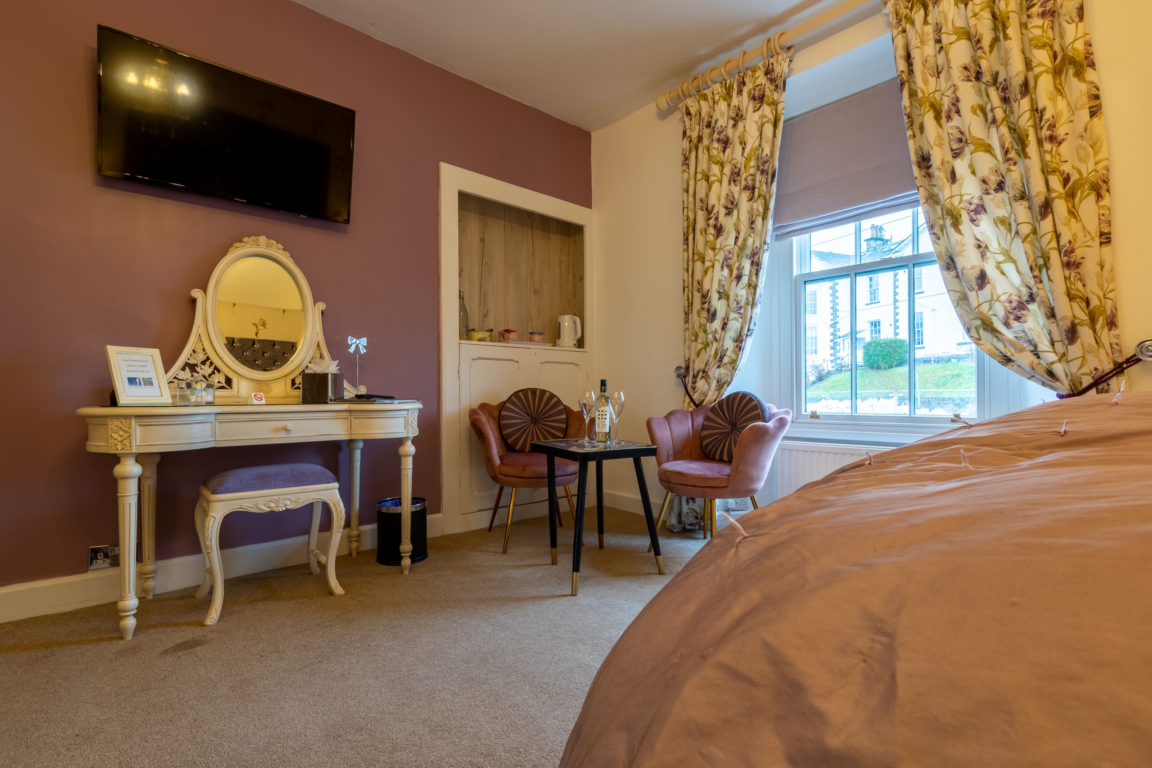 A bedroom with a bed, a desk with a mirror, two chairs, a small table with a lamp, and a window with curtains.