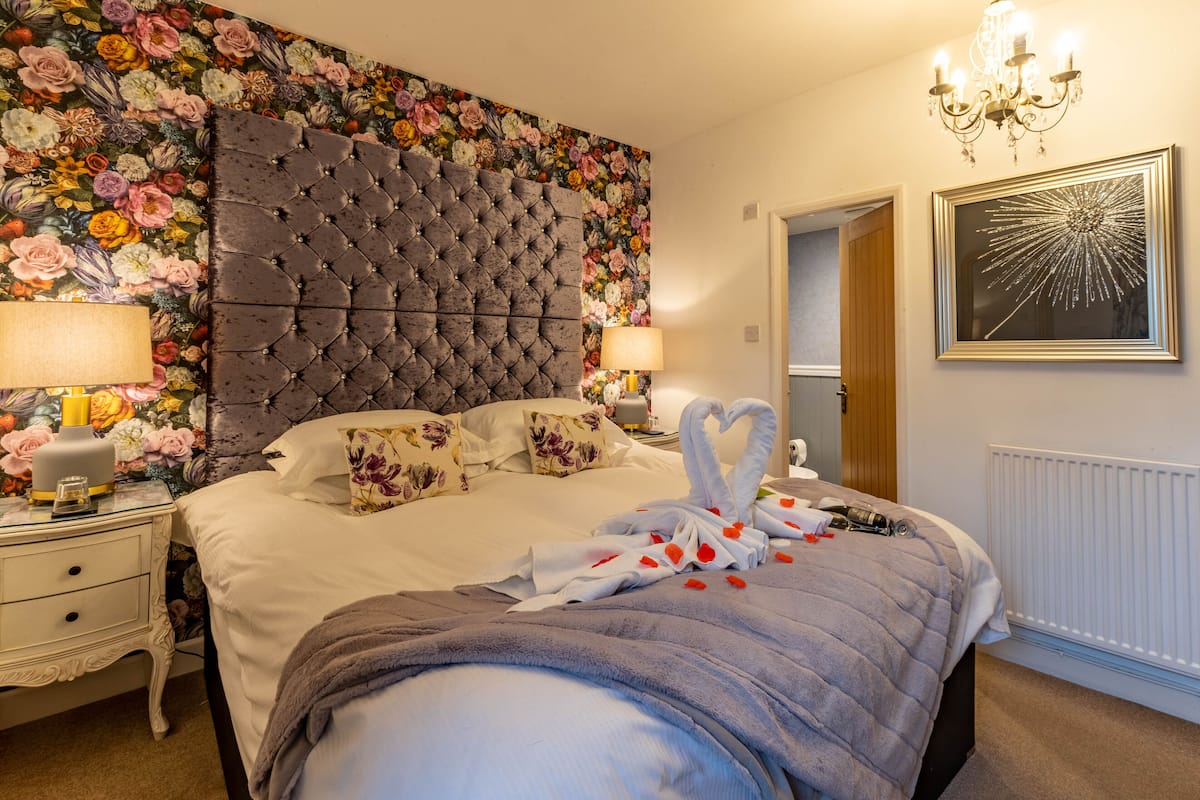 A bedroom with a tufted headboard, floral wallpaper, a bed with a grey blanket, two bedside tables with lamps, and a chandelier.