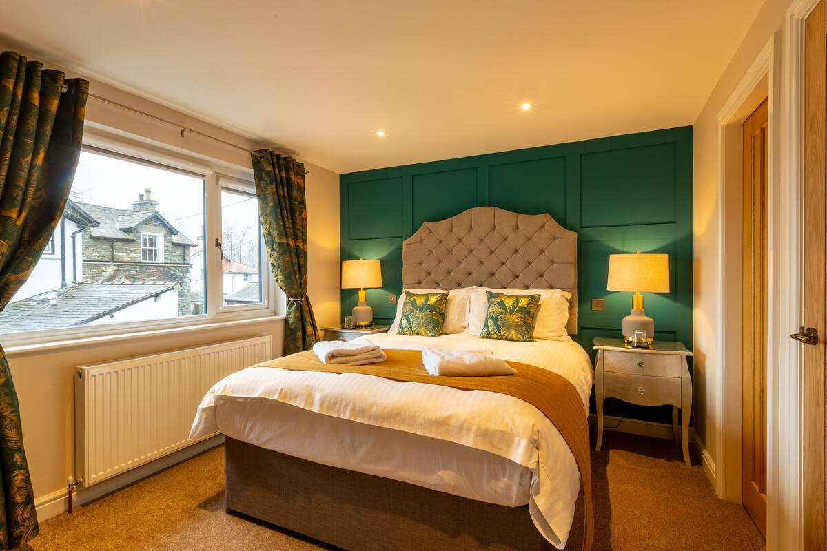 A hotel room with a large bed, two bedside tables with lamps, a view of a house through the window, and teal-colored walls.