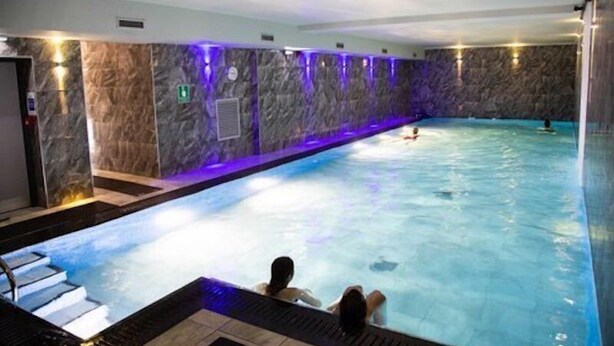 An indoor swimming pool with a marble wall, a person relaxing in the water, and another person swimming. - Holly Lodge Guest House (Windermere)