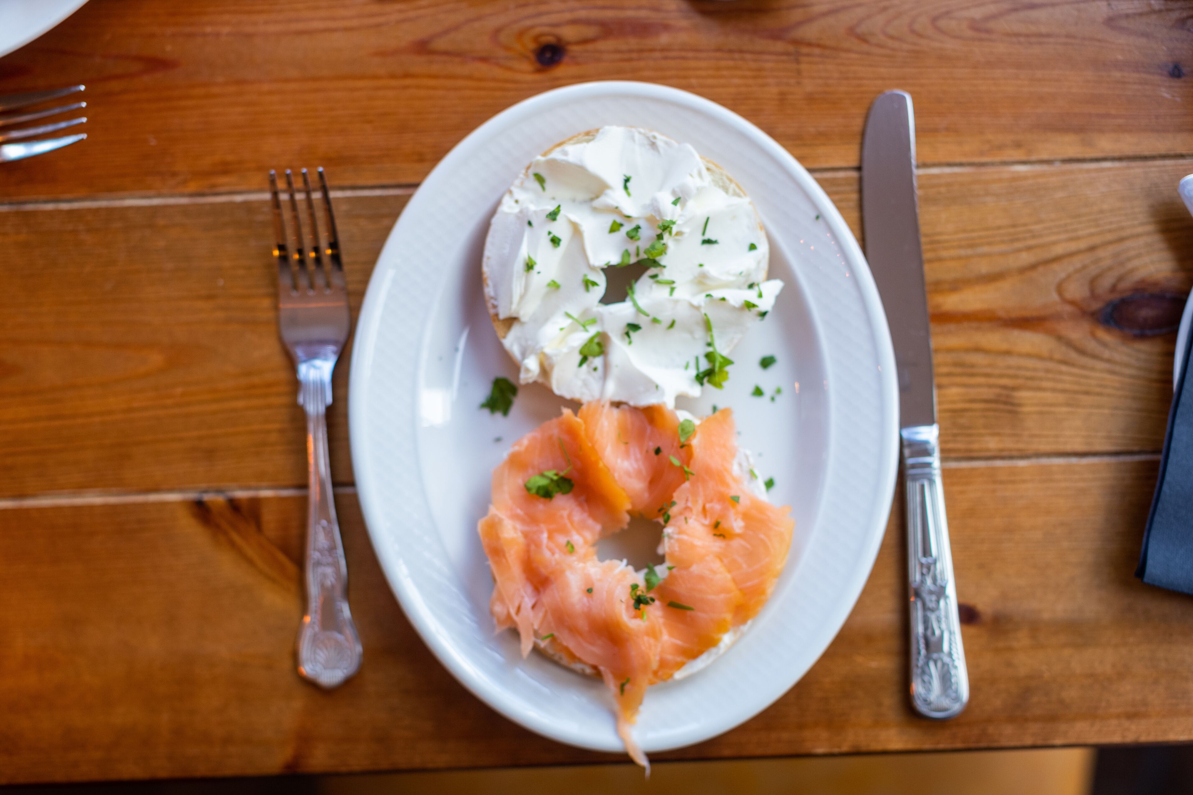 A plate with a bagel topped with cream cheese and salmon, accompanied by a fork and knife on a wooden table.