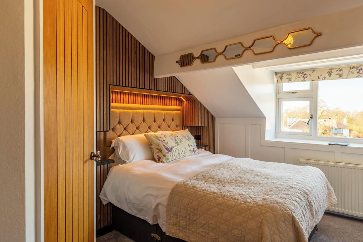 A bedroom with a sloped ceiling, a bed with a quilted bedspread, a wooden headboard, and a window with a view of houses.