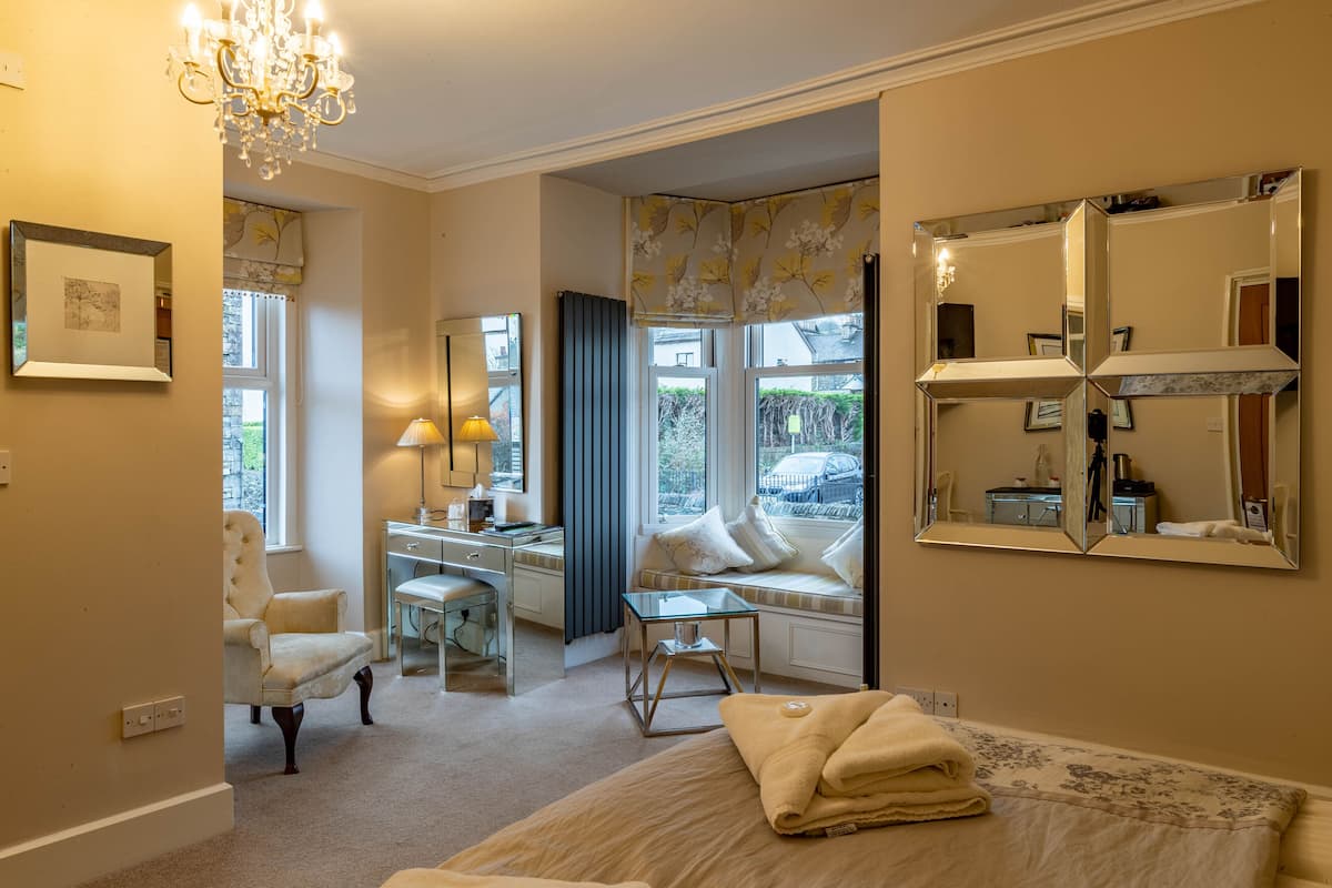 A bedroom with a bed, a chair, a desk, a lamp, a window with a view, and a large mirror.