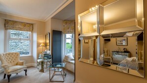 A room with a large mirror, a plush armchair, a glass table, and a view of the outdoors through a window with a stone facade. - Holly Lodge Guest House (Windermere)