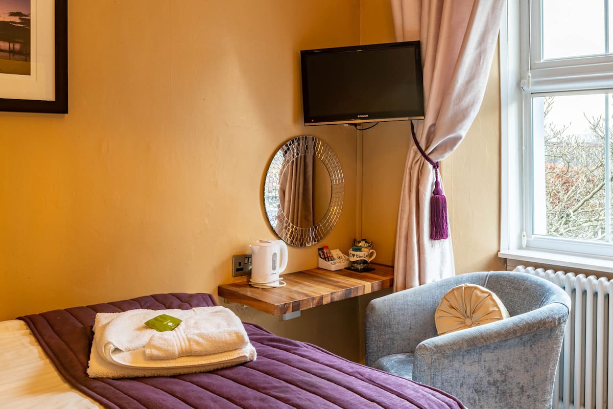 A hotel room with a bed, a chair, a TV, a mirror, a window, and a small table with a kettle and a mug.