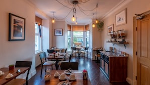 A dining area with wooden tables and chairs, a kitchen area with a wooden counter, and a view through the bay window. - Holly Lodge Guest House (Windermere)