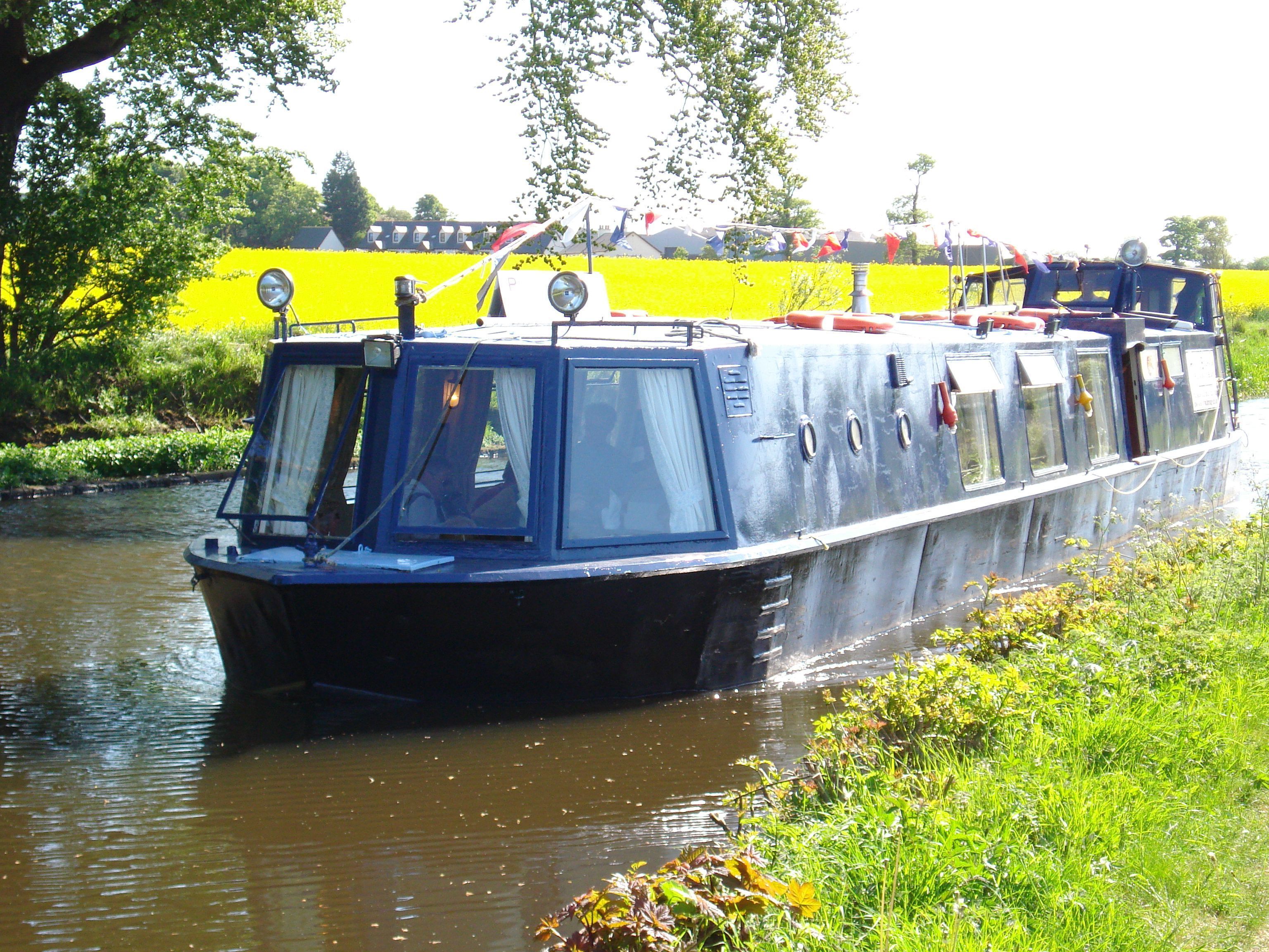 A narrowboat with a flat roof and windows, moving through a canal with greenery on the banks.