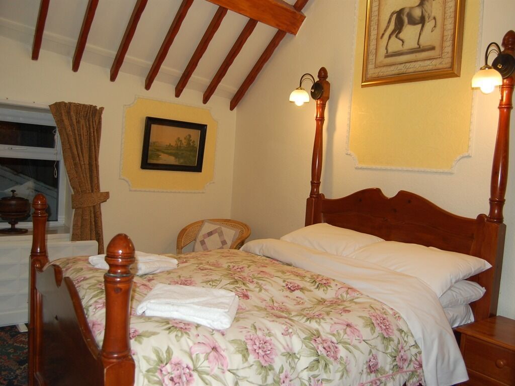 A bedroom with a wooden four-poster bed, a nightstand, a chair, a window with curtains, and a framed picture on the wall.
