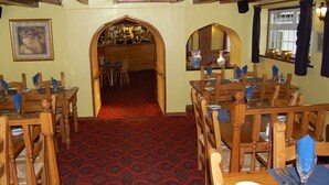 A restaurant with wooden tables and chairs, a red patterned carpet, and a bar area visible through an archway.