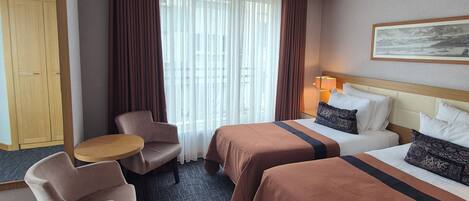 A hotel room with two beds, a desk, a chair, a window with curtains, and a painting on the wall.