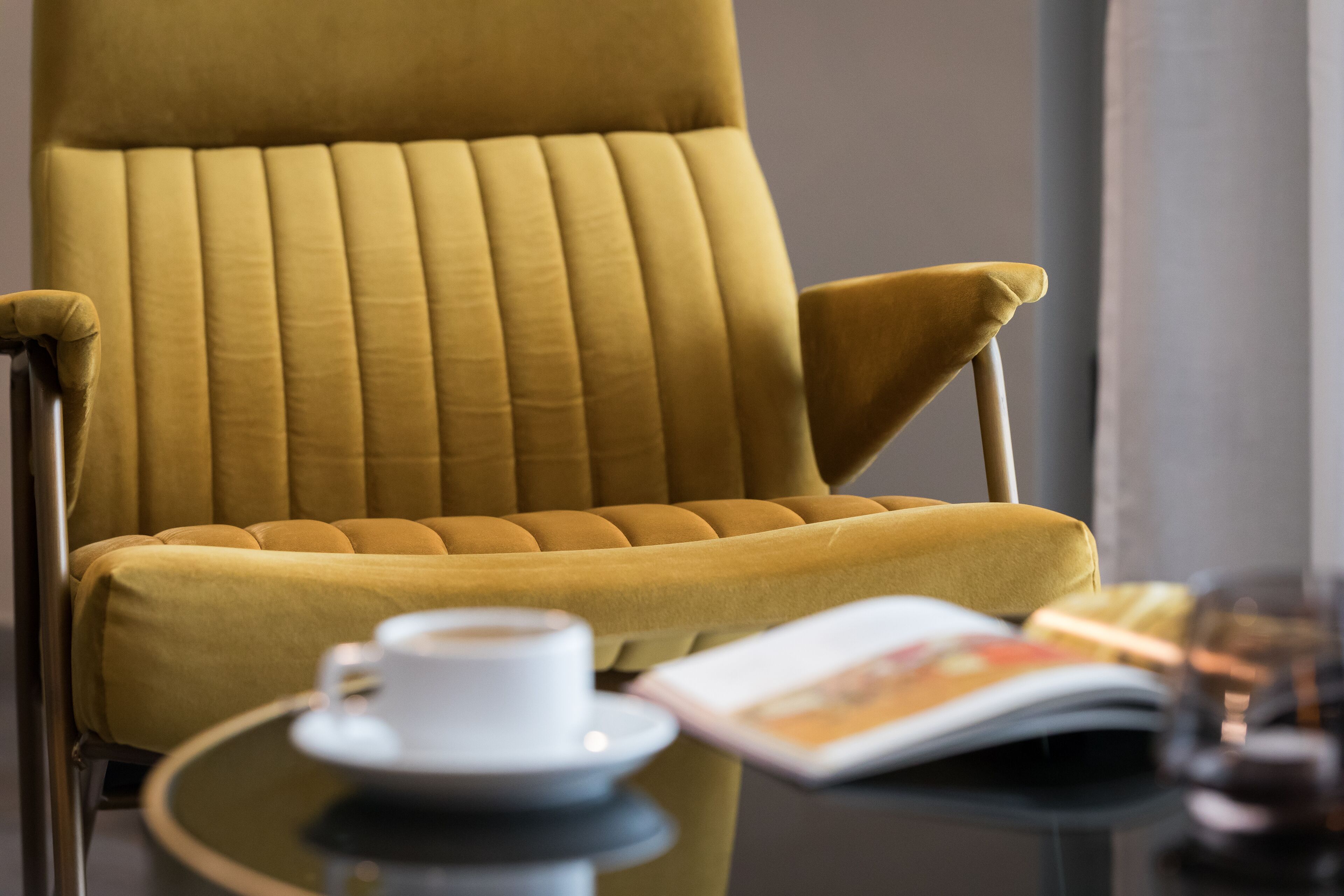 Un sillón de color mostaza con asiento y respaldo acolchados, ubicado junto a una mesita redonda con una taza y platillo blancos, y una revista.