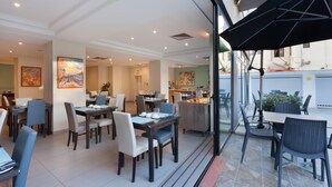 Un comedor moderno con mesas y sillas, una cocina al fondo y vistas a través del muro de cristal.