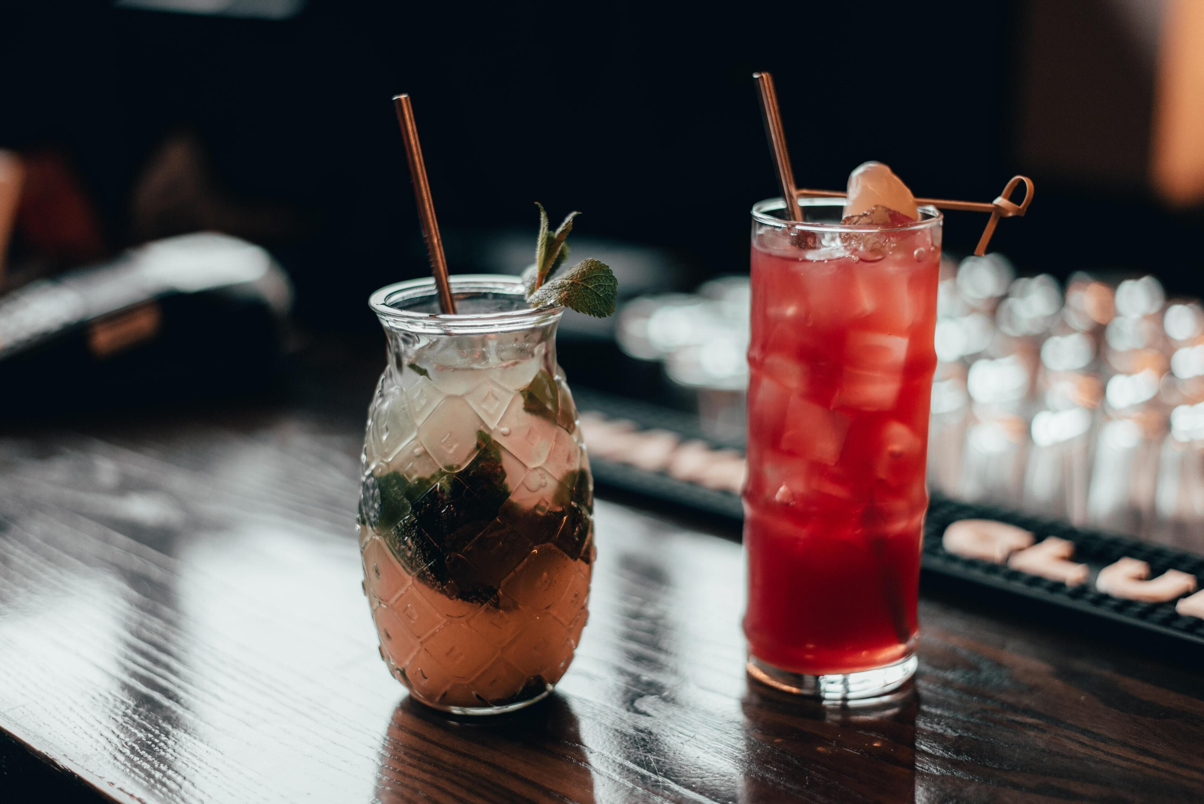 Two drinks on a bar counter, one in a mason jar and one in a tall glass, with straws and a garnish.