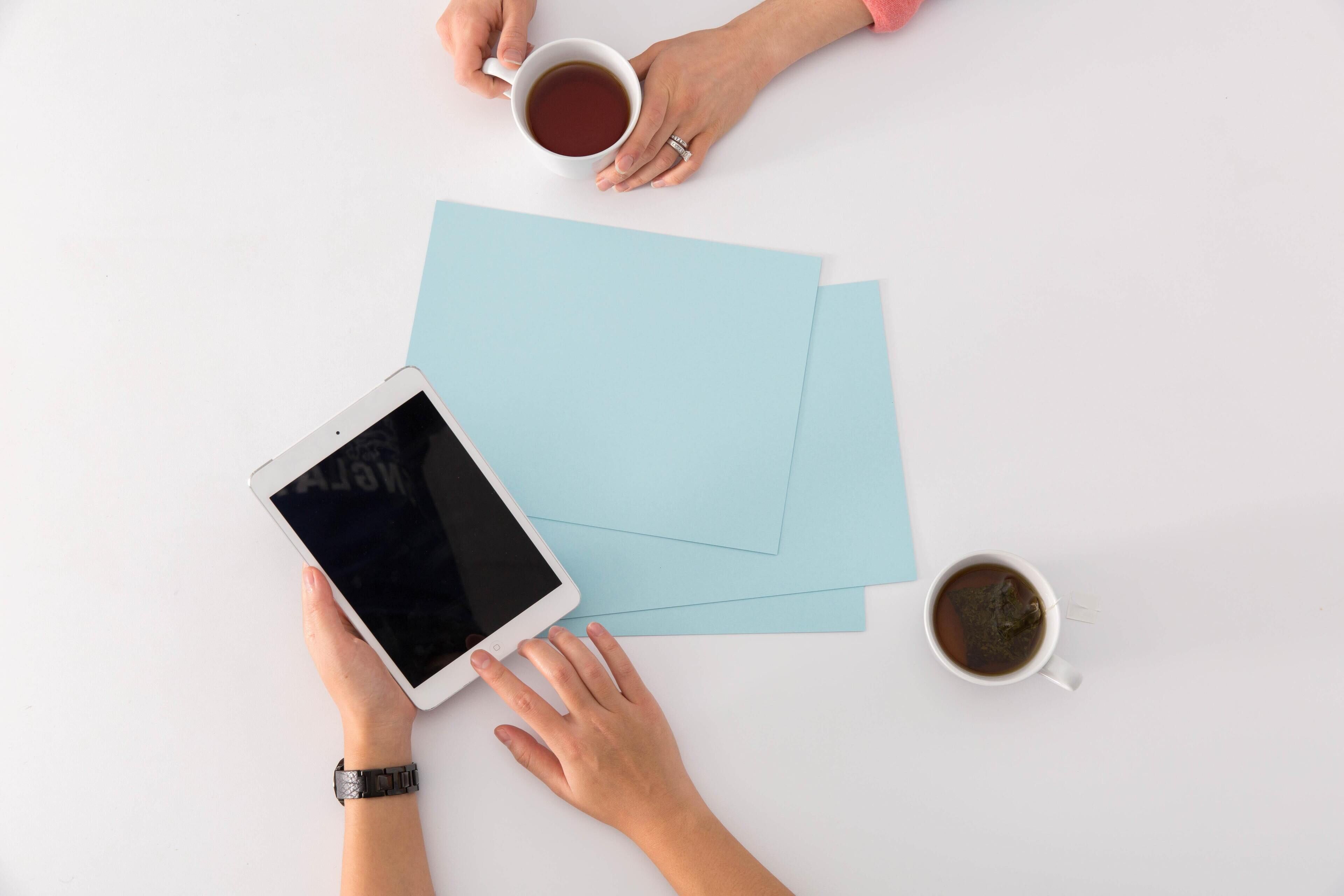 A white table with a tablet, two cups of coffee, and two blue sheets of paper.