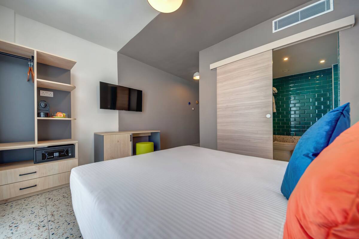 A modern hotel room with a large bed, built-in TV, and a shower area with green tiles.