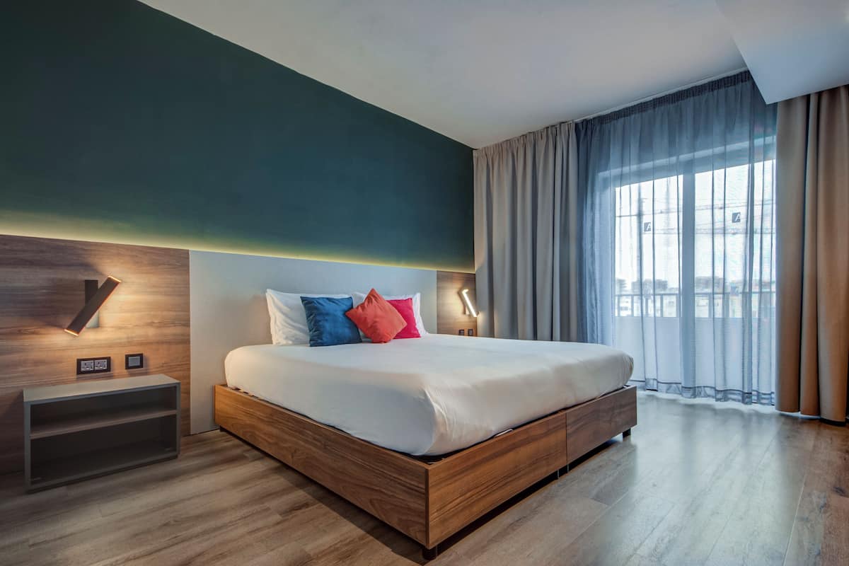 A modern hotel room with a large bed, wooden headboard, bedside tables, and a view of the city through large windows.