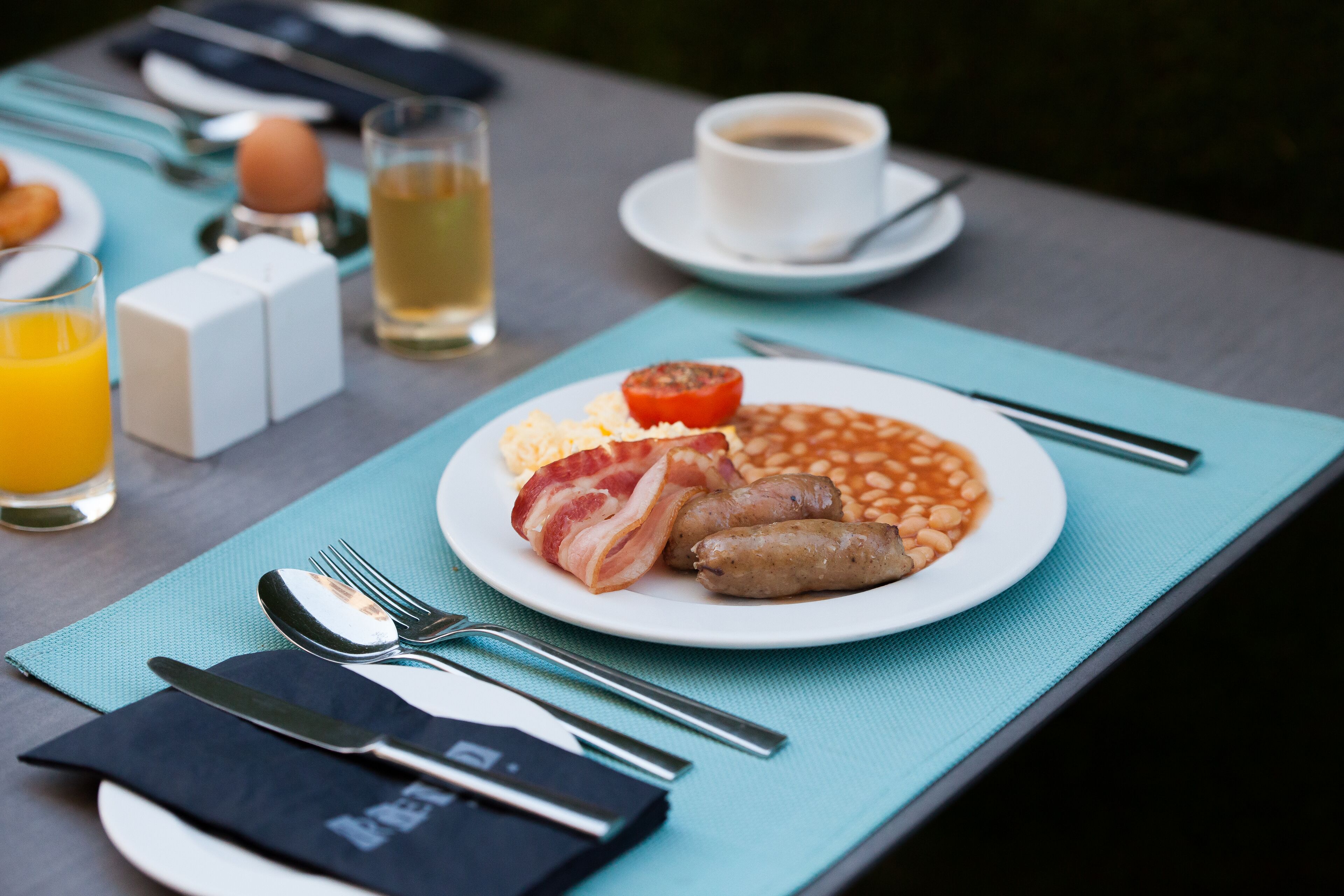 Ein Frühstückstisch mit Bohnen, Würstchen und Speck, einem Glas Orangensaft und einer Tasse Kaffee.
